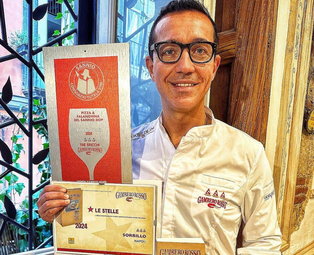 This photo shows a bespectacled Gino Sorbino in a white chef's coat holding up various awards he has won for his pizza-making skills.