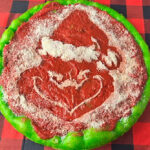 This photo shows a round Philly Tomato Pie pizza with a grinning Grinch in the center and green dough around the edges.