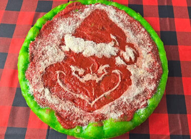 This photo shows a round Philly Tomato Pie pizza with a grinning Grinch in the center and green dough around the edges.