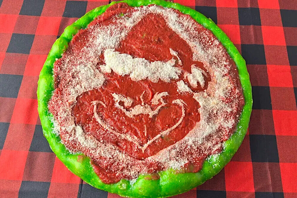 This photo shows a round Philly Tomato Pie pizza with a grinning Grinch in the center and green dough around the edges.