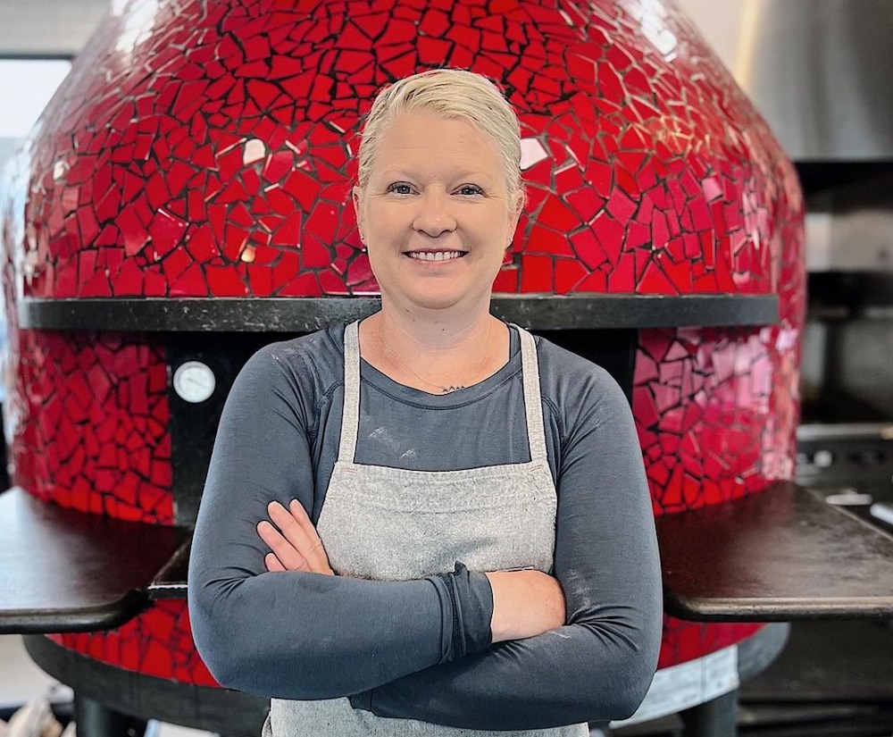 This photo shows Kerri Smith, with her arms crossed, standing in front of a red wood-fired pizza oven and smiling.