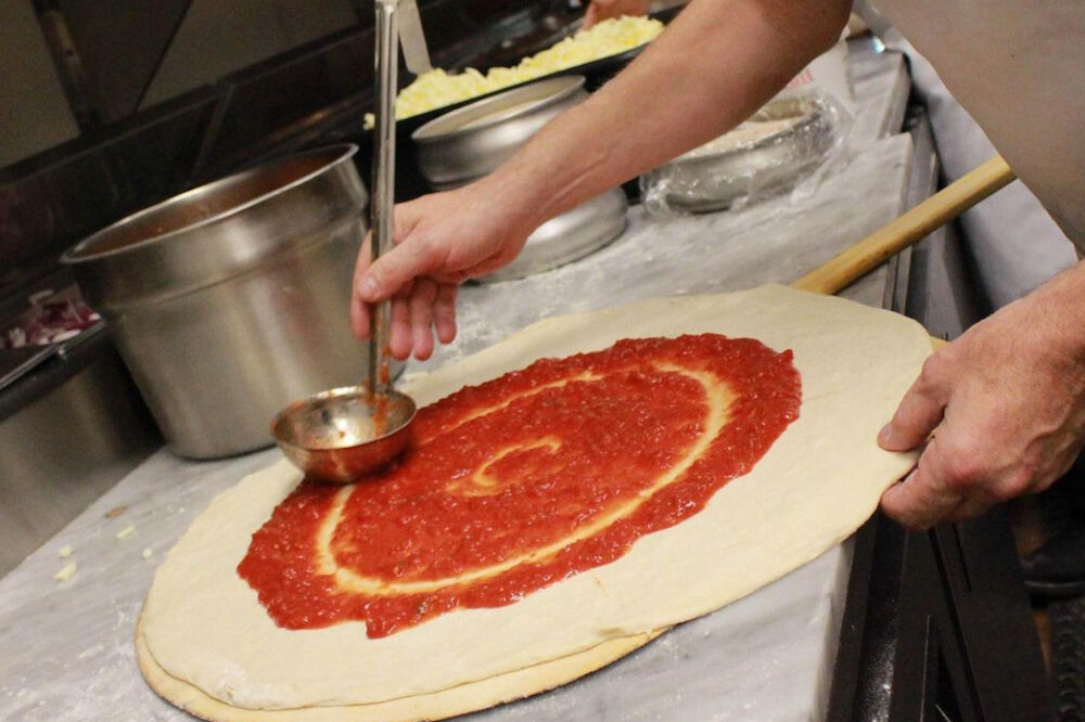 This photo shows a pizza maker (from the elbow down) spreading sauce on a New York-style pizza crust.