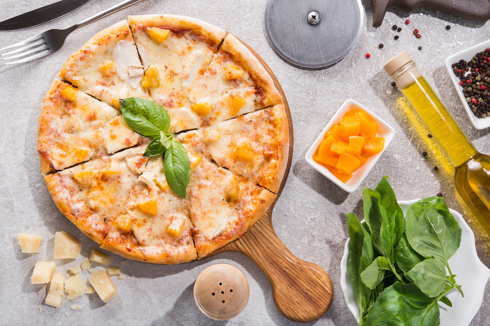 This photo shows a pizza topped with chicken fillets and peaches, with other ingredients, including a bowl of diced peaches to the right side.