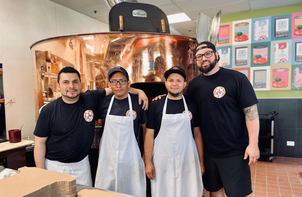 This photo shows a bearded, capped Ric Gruber standing with three male members of his staff in front of their pizza oven.
