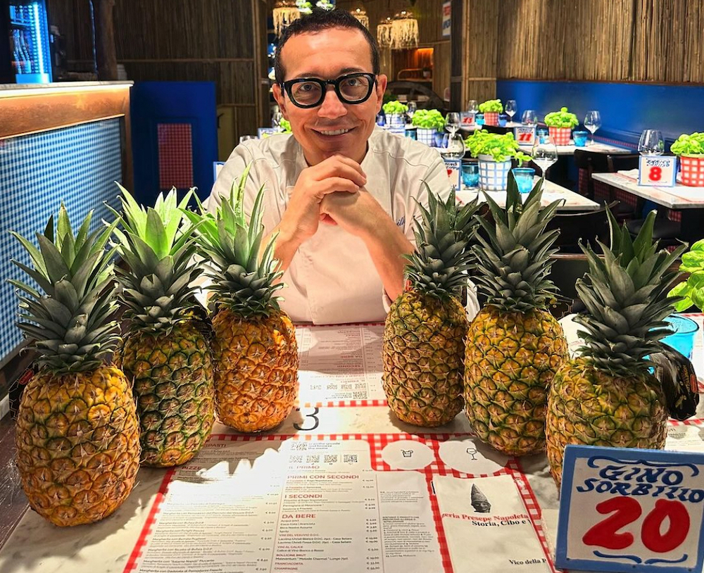 This photo shows Gino Sorbillo with his chin resting on his hands, sitting at a table arrayed with six whole raw pineapples.