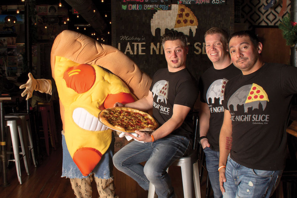 This photo shows the three owners of Mikey's standing together, with PizzaFace, their mascot, standing at far left. PizzaFace is a human-sized pepperoni pizza slice.