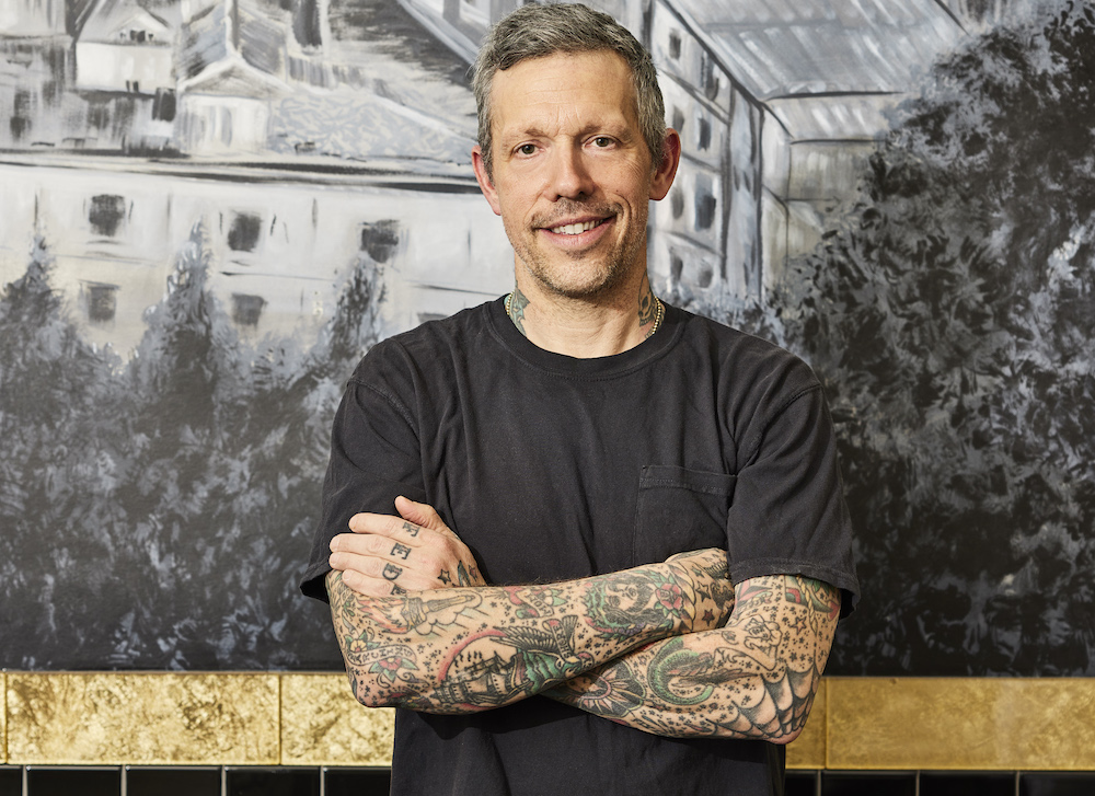 This photo shows Anthony Mangieri, with arms crossed and wearing a black t-shirt, standing in front of a mural at Una Pizza Napoletana