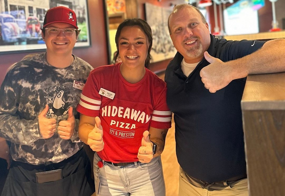 This photo shows a young woman in the center wearing a red Hideaway Pizza shirt, in between a younger man in a red cap and an older bearded man in a black shirt, all giving the thumbs-up sign.