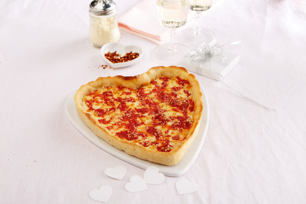 This photo shows a heart-shaped cheese pizza against a white table cloth set with a small white container for red pepper flakes and a Parmesan cheese shaker.