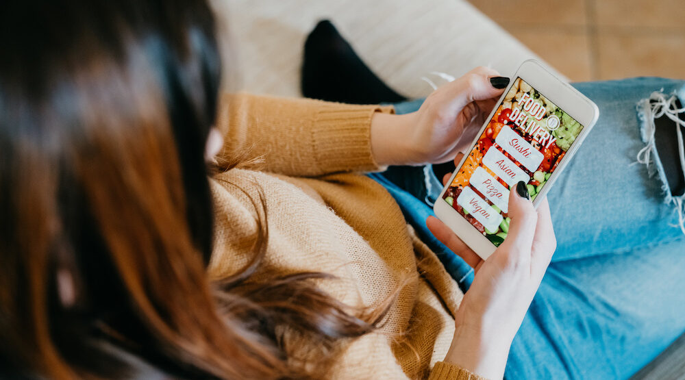 This photo shows a woman with long brown hair, shown from behind and over her shoulder, holding a smartphone with various food ordering options.