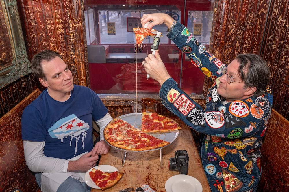 Two men sit at a booth at John's of Bleecker Street, playfully testing the audio levels of a cheese of pepperoni pizza. The men are podcasters.