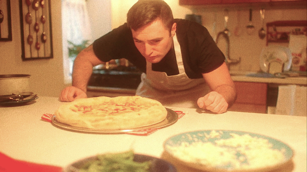 This vintage photo shows a young Anthony Mongiello leaning in and peering closely at a stuffed crust pizza that he has made himself.