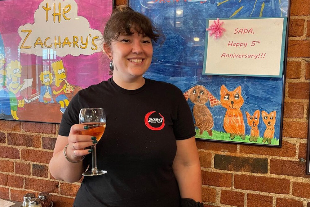 This photo shows a young woman in a black Zachary's t-shirt holding a glass of wine