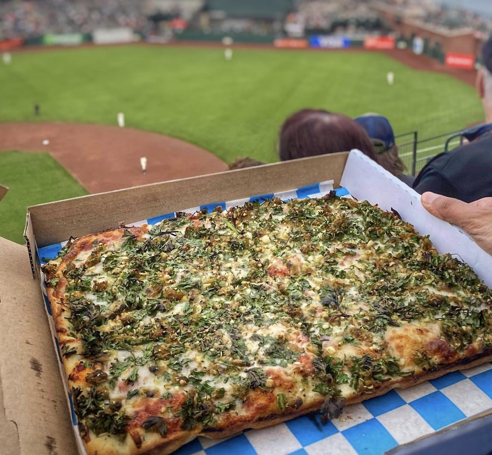 A signature Golden Boy pizza sits in a box with the backdrop of a San Francisco Giants game. The person eating the pizza and taking the photo are attending a live baseball game.