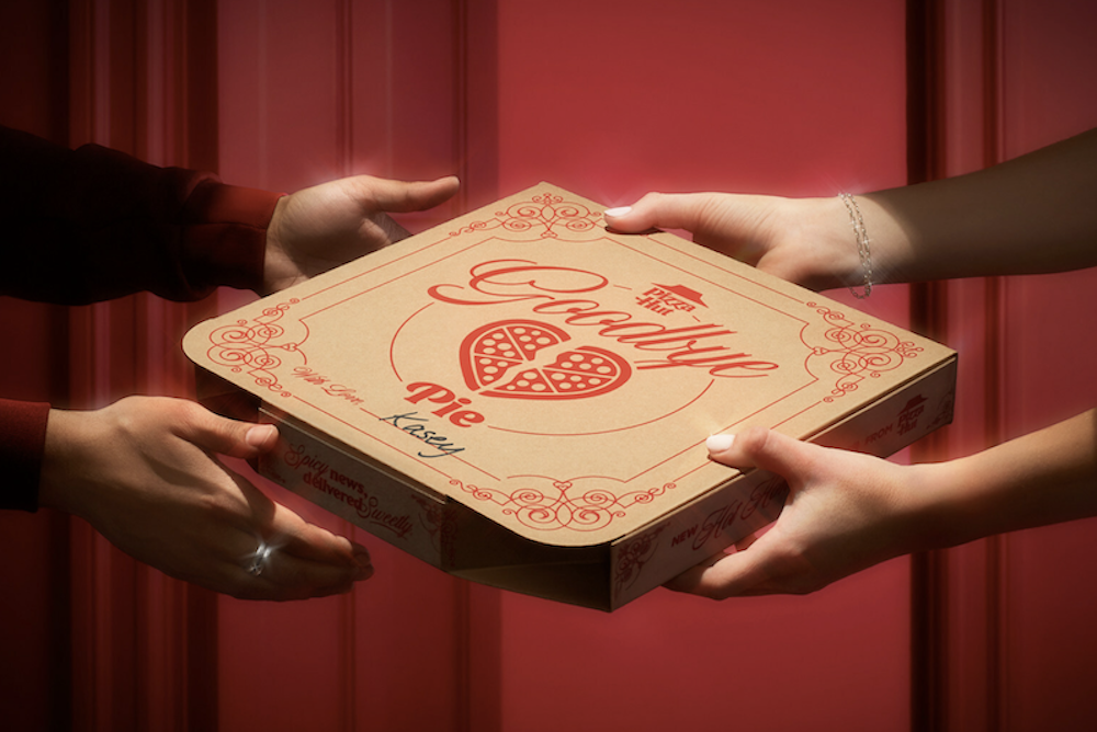 One person's set of hands gives a pizza to someone else's awaiting set of hands. Inscribed on the pizza box is the name "Kasey."