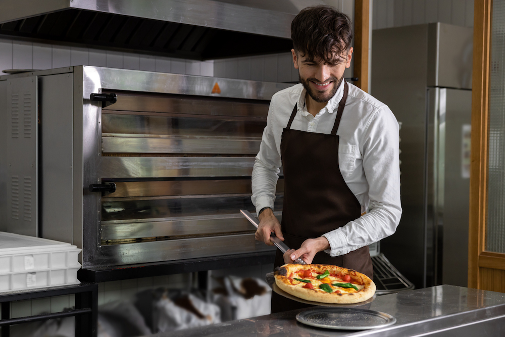 This photo shows a bearded man in a white shirt and black apron with a fresh pizza removed from a set of deck ovens behind him.