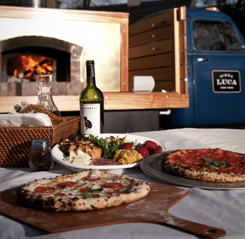 A spread of pizza, wine and flowers sits in the foreground with a pizza truck and wood-fired pizza oven in the background.