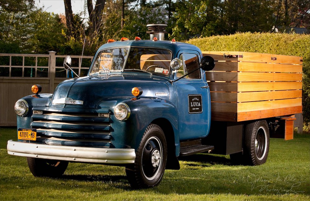 A blue 1952 Chevy flatbed with a pizza oven mounted on the back. A graphic reads "Pizza Luca" on the driver's side door.