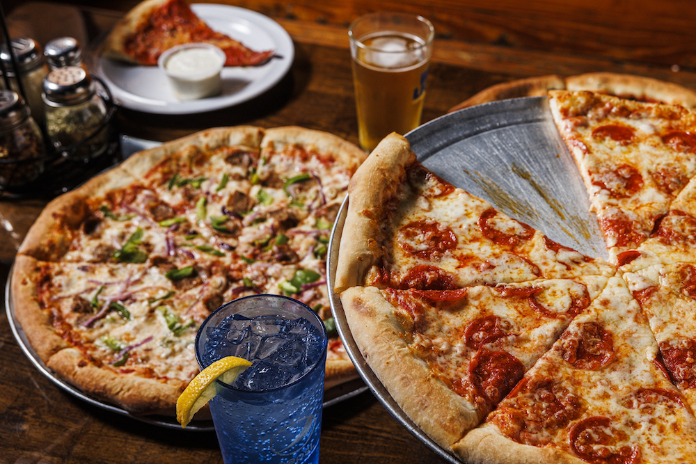 This photo shows two different thin-crust pizzas topped with ingredients like pepperoni, sausage and bell peppers from Village Idiot Pizza.