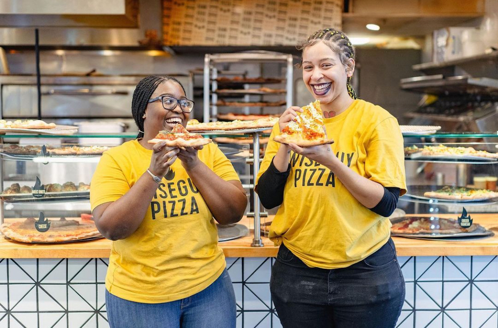 This photo shows two young women in yellow Wiseguy Pizza t-shirts smiling and enjoying slices.