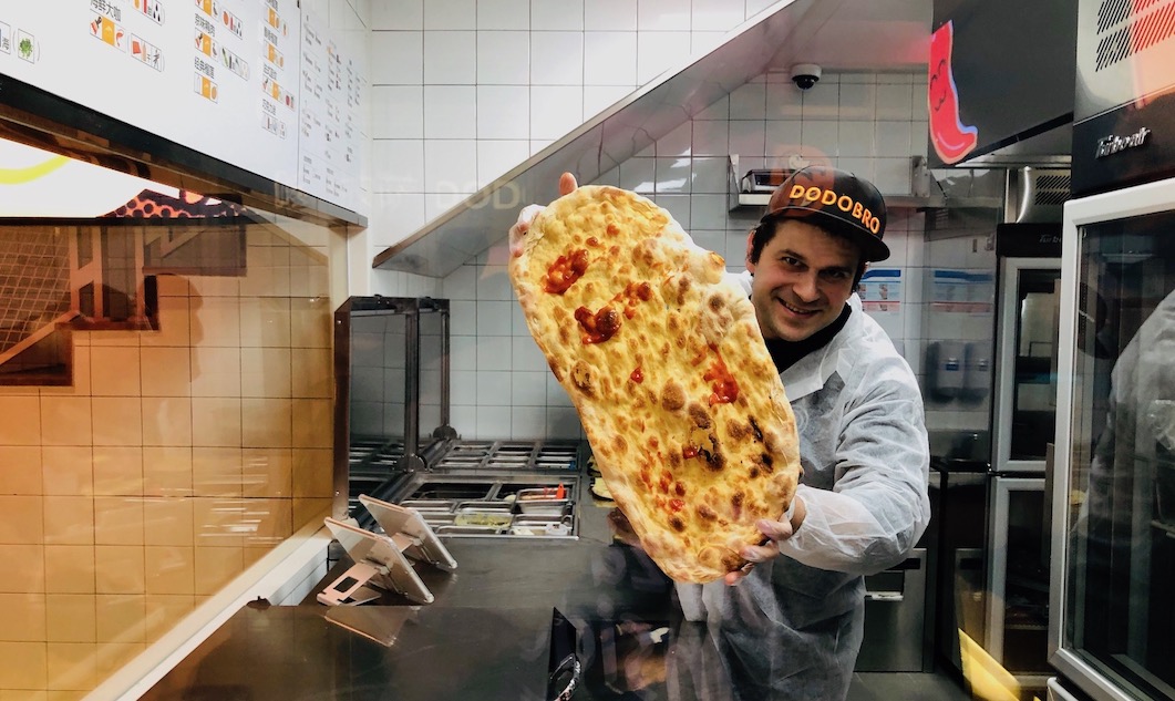 This photo shows a male Dodo Pizza employee holding an oblong-shaped pizza in the kitchen.