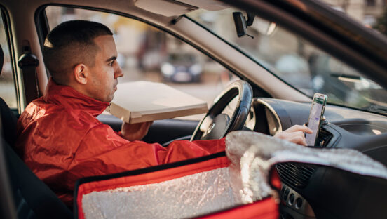 This photo shows a male delivery driver with close-cropped hair checking a phone mounted on the dashboard of his car.