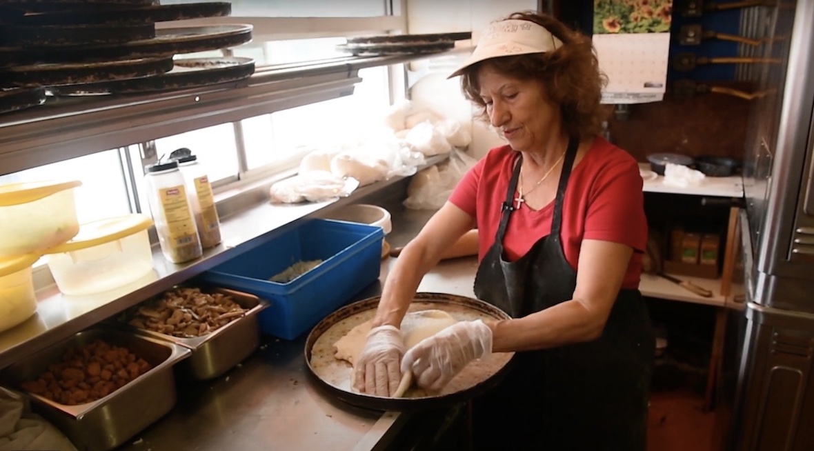 This photo shows Elli Darmoyslis stretching pizza dough in a pan at The Varsity. She's wearing a red shirt, a black apron and a white visor.