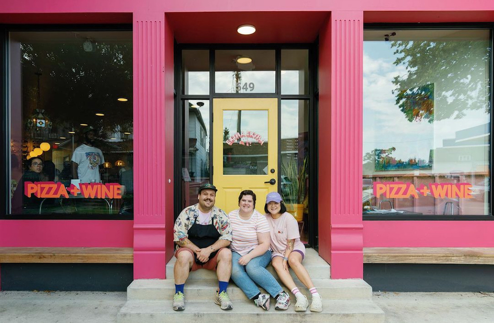 The photo shows the three owners, casually dressed, sitting on the steps of the front entrance to their pink-colored pizzeria
