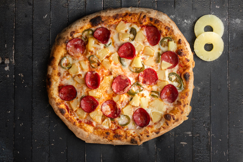 This photo shows a Hawaiian pizza with pineapples and pepperoni along with two round pineapple slices arranged into the figure 8