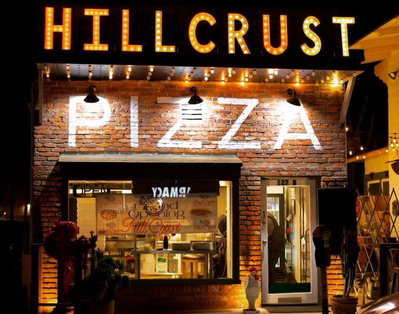 This photo shows the brightly lit exterior of Hillcrust Pizza as seen at night, with its neon signs lit up.