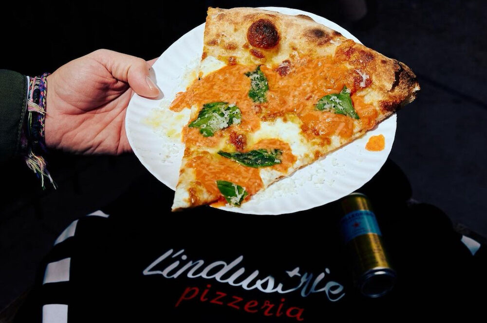 This photo shows a slice of a vodka pizza with fresh mozzarella set against a black L'Industrie t-shirt