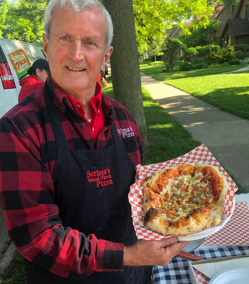 Joe Scrima, wearing a red and black plaid shirt, displays one of his pizzas.