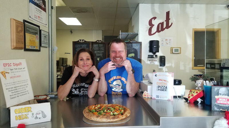 This photo shows Kira and Mark, with their chins eating on their hands, smiling at the service counter with a pizza in front of them.