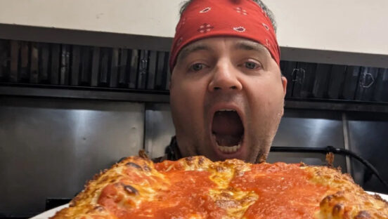 This photo shows Matty Evans, wearing a red banana, with his mouth wide open as he gets ready to bite down a whole Detroit-style pizza.