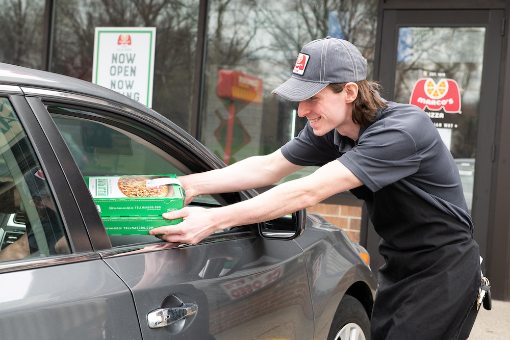 A Marco's Pizza employee hands a pie through the driver-side window of a car.