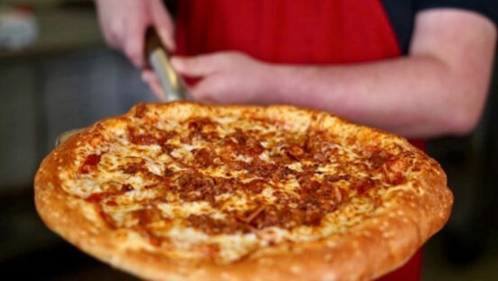 This photo shows a man's hands holding a pizza peel with a cheese pizza.