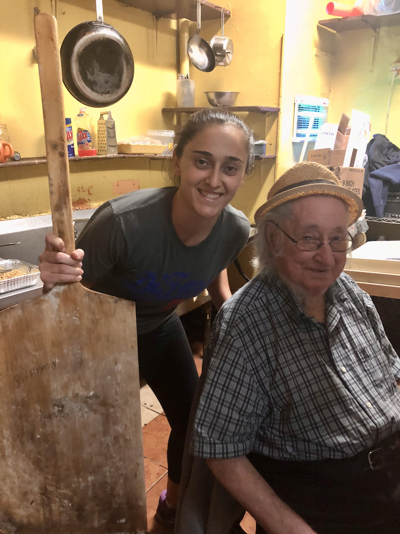 This photo shows Shannon holding a wooden pizza peel and bending over to be photographed with the legendary pizza maker Dom DeMarco.