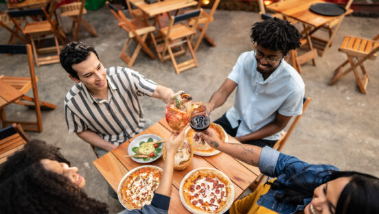 This photo shows a diverse group putting their beer and wine glasses together in a toast at a table with pizzas.