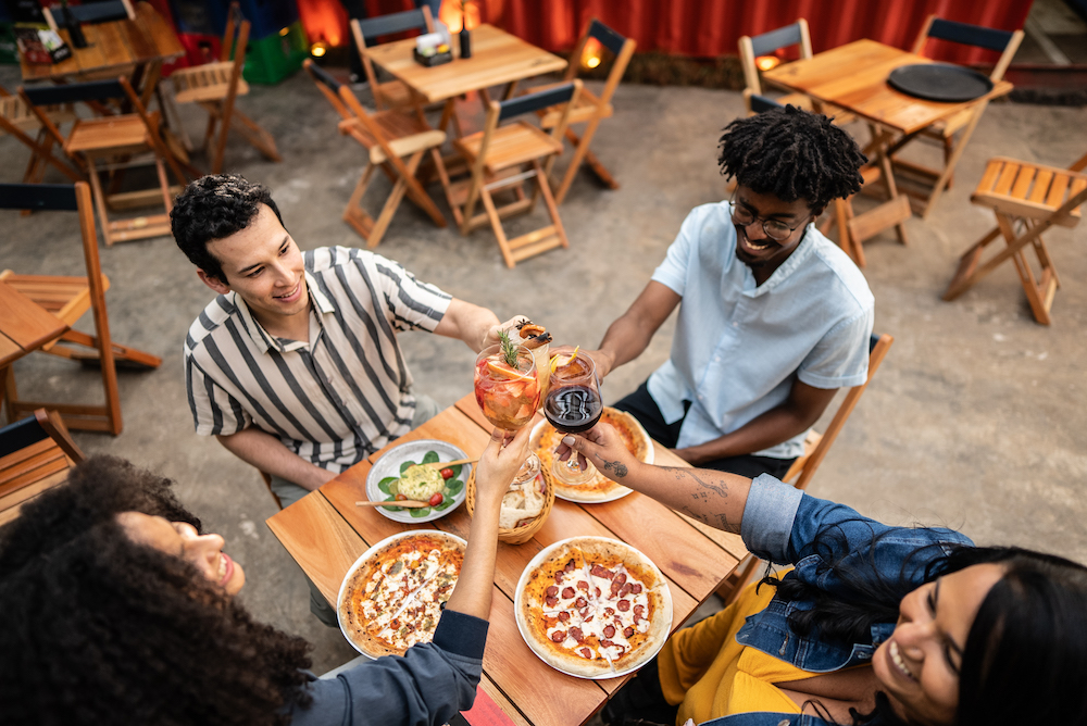 This photo shows a diverse group putting their beer and wine glasses together in a toast at a table with pizzas.