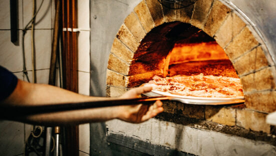 This photo shows a man putting a pizza in a wood-fired oven.