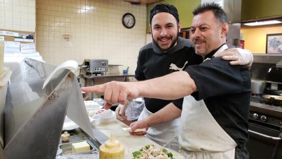 This photo shows Andrea Gramo in a black chef coat and a white apron, pointing to his POS system while an employee in a black shirt looks on, with both men smiling.