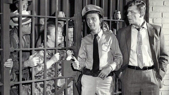 This photo shows a scene from The Andy Griffith Show, with Barney Fife, in uniform, and Andy Taylor, in a suit, standing in front of a jail cell that's packed with local citizens arrested by Barney, including Aunt Bee.