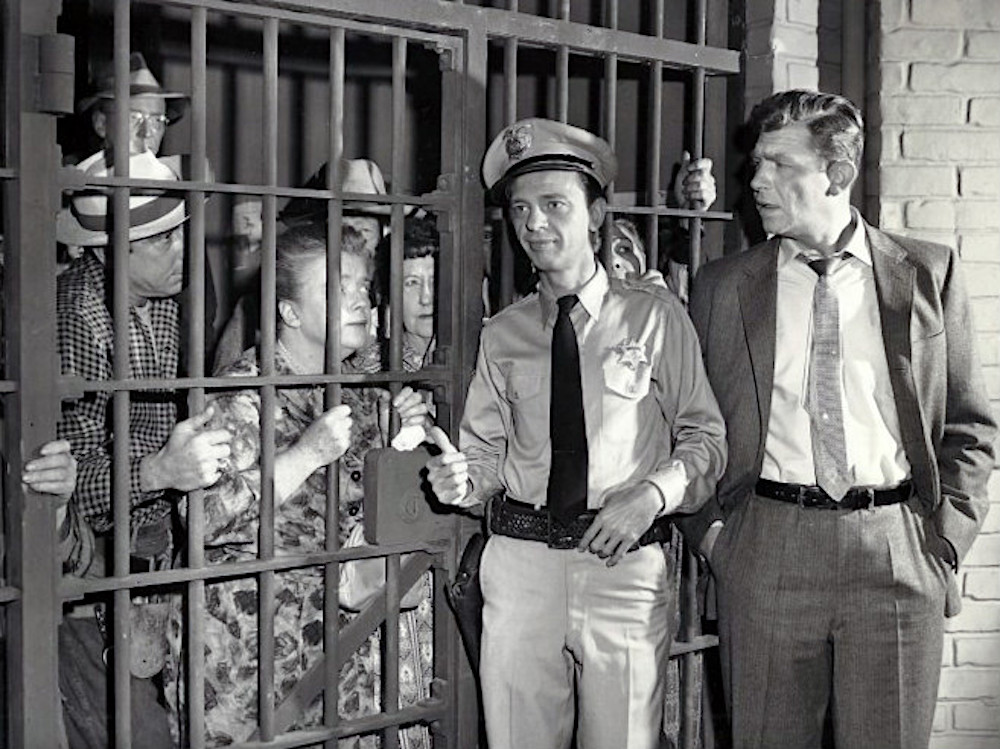 This photo shows a scene from The Andy Griffith Show, with Barney Fife, in uniform, and Andy Taylor, in a suit, standing in front of a jail cell that's packed with local citizens arrested by Barney, including Aunt Bee.