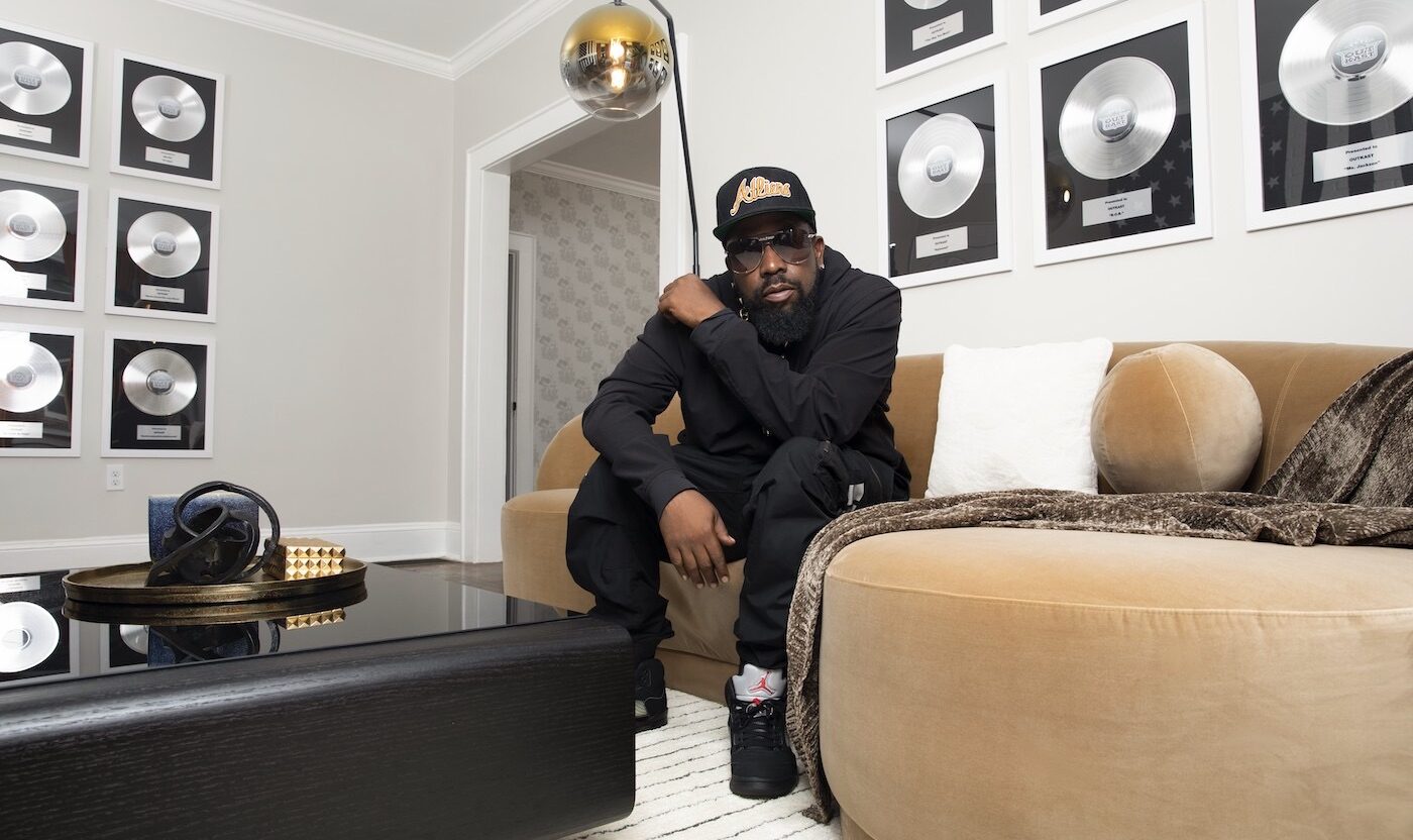 This photo shows rapper Big Boi, dressed all in black, sitting on a sofa in a room surrounded by what appear to be awards for platinum-selling records.