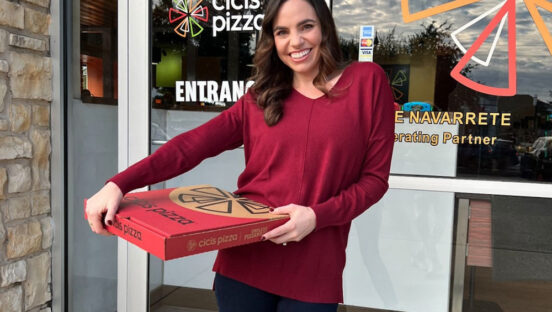 This photo shows a smiling young woman with long dark hair and a red sweater exiting a Cicis Pizza restaurant with a box of pizza.