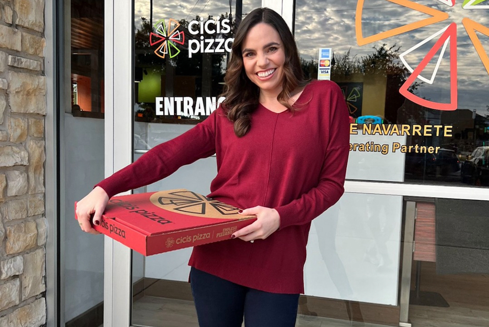 This photo shows a smiling young woman with long dark hair and a red sweater exiting a Cicis Pizza restaurant with a box of pizza.