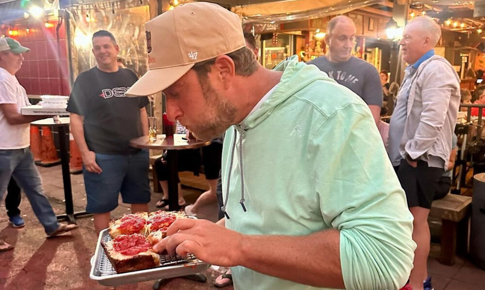 This photo shows Dave Portnoy in a light green sweatshirt trying a Detroit-style pizza slice at The Nona Slice House.