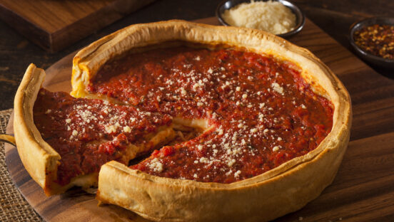 This photo shows a whole deep-dish pizza with one slice pulled slightly away from the whole against a wooden background.