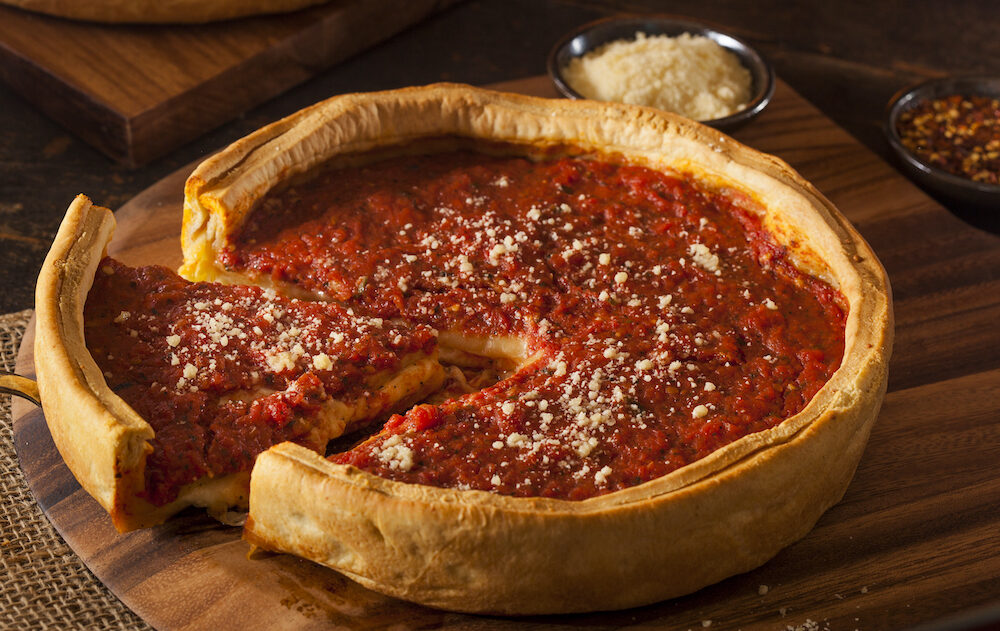 This photo shows a whole deep-dish pizza with one slice pulled slightly away from the whole against a wooden background.