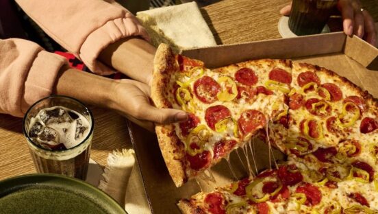 This photo shows a woman's hand holding a slice of New York-style pizza removed from a whole pie in a carryout box.